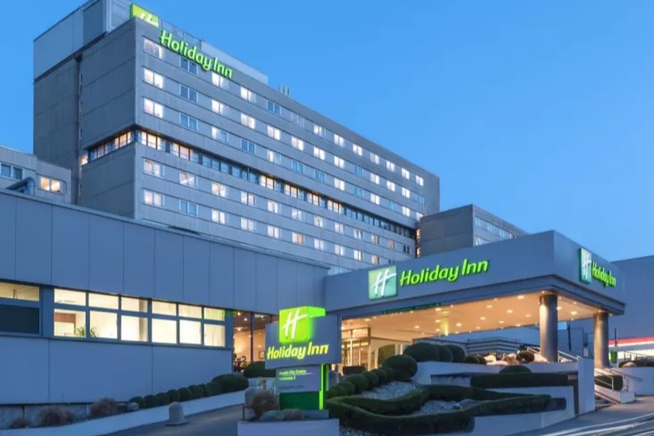 Image of a modern Holiday Inn hotel exterior during the evening, adorned with bright green and white signage. The entrance is well-lit and features a covered area with decorative landscaping reminiscent of Old World charm, evoking the festive spirit found in Christmas markets nearby.