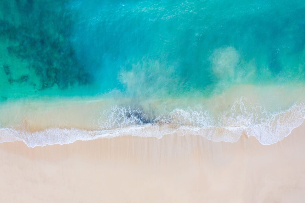 Aerial view of a tranquil Bali beach with soft, white sand meeting clear, turquoise water. Gentle waves create a lacy edge along the shoreline. The gradient of blue in the water varies from deep to light, reflecting the natural beauty of the ocean.