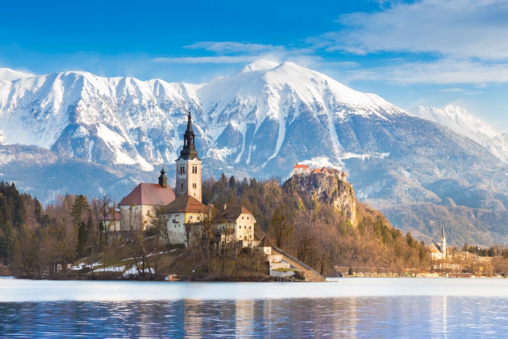 A serene lakeside scene with a church and castle on an island, surrounded by calm waters. Snow-capped mountains and blue skies with scattered clouds form a majestic backdrop. The landscape exudes tranquility and Old World natural beauty, reminiscent of scenes near traditional Christmas Markets.