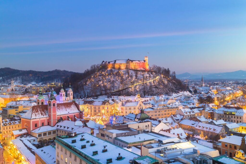 A snowy winter evening in Ljubljana, Slovenia, featuring a beautifully illuminated Ljubljana Castle atop a hill. The historic cityscape below showcases warmly lit buildings and streets adorned with festive Christmas lights, blending classic Old World architecture with seasonal cheer.