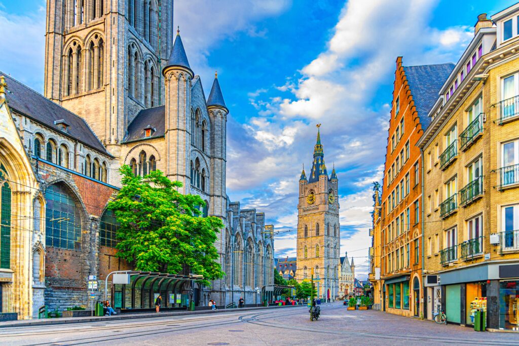 A vibrant street scene in Ghent, Belgium, showcases St. Nicholas' Church on the left with its historic gothic architecture. Cobblestone streets lead towards the Belfry of Ghent in the background. Modern buildings and shops line the right side under a bright sky reminiscent of an Amsterdam summer day.