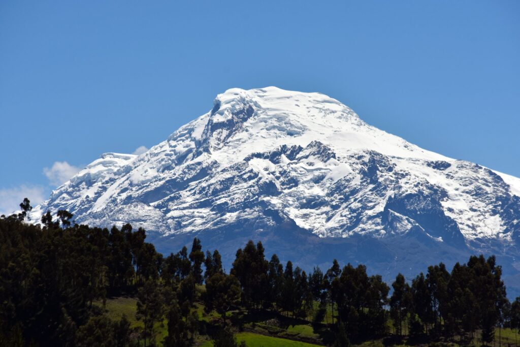 A snow-capped mountain peak rises majestically against a clear blue sky in Ecuador. Below, lush green trees and vegetation line the lower slopes, contrasting with the white of the snow. The scene is serene and picturesque, reminiscent of a view you might encounter on a Galapagos Islands tour.