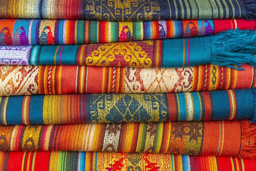 A stack of vibrant, multicolored woven textiles with intricate patterns and designs evokes the rich culture of an Ecuador and Galapagos Islands tour. The fabrics display a variety of stripes, geometric shapes, and traditional motifs in hues of red, blue, green, orange, and yellow.