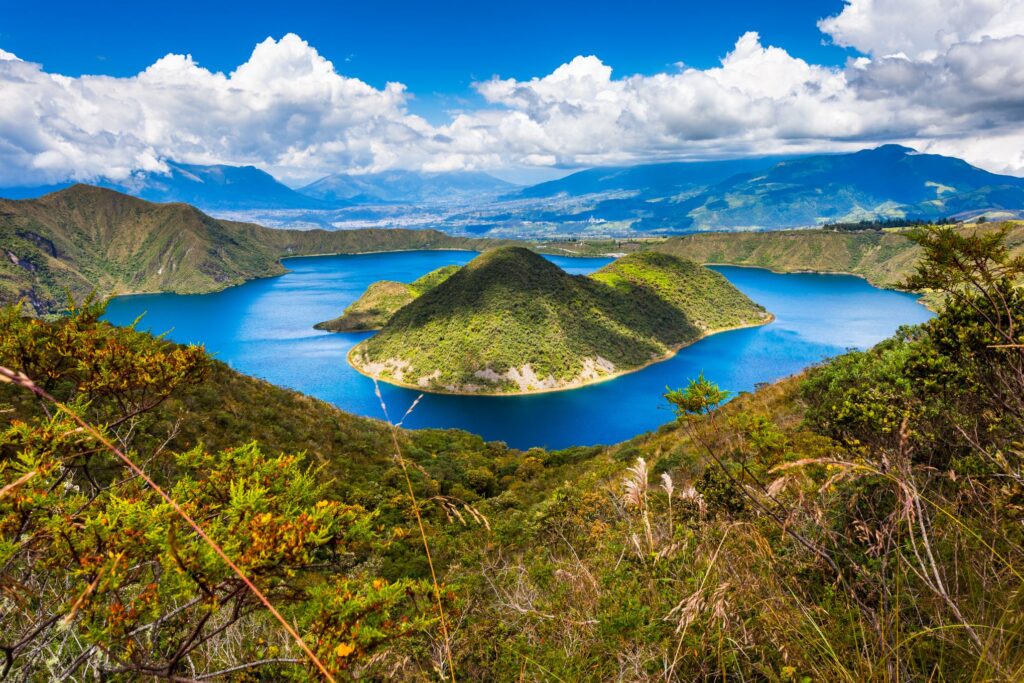 Scenic view of a tranquil blue lake surrounded by lush green hills and mountains under a partly cloudy sky. The landscape includes a few small vegetated islands within the lake, reminiscent of an Ecuador and Galapagos Islands tour, with distant mountain ranges in the background. Dense foliage is in the foreground.