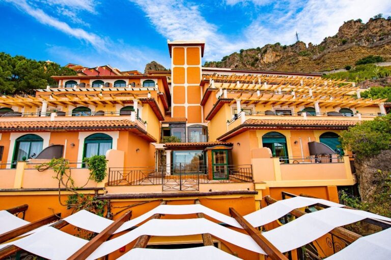 A vibrant, multi-story building with a Mediterranean architectural style and a bright orange facade features several balconies and a large terrace. Pergolas with white fabric shades extend from the building, surrounded by lush greenery and hills, evoking the rich flavors of Sicily.