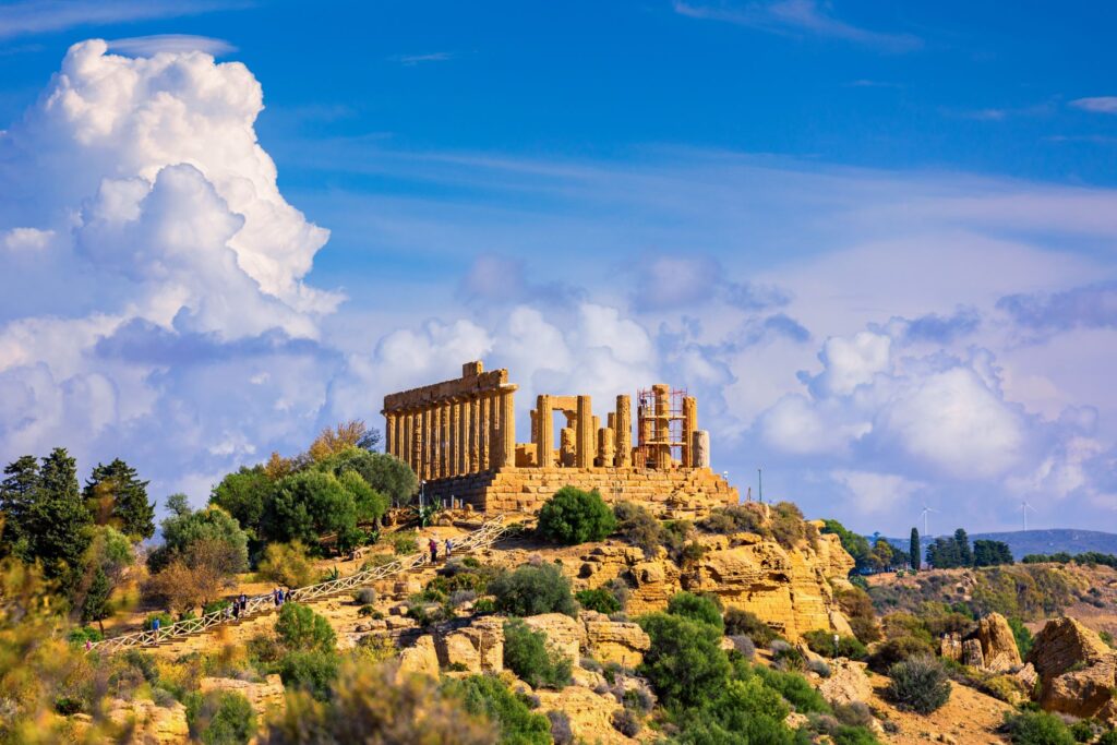 Ancient Greek Doric temple ruins, set on a rocky hilltop amidst greenery and under a partly cloudy blue sky. The scene includes iconic columns and scattered stone remnants, evoking a sense of historical grandeur and natural beauty, much like the rich Sicilian flavors that define the island's cuisine.