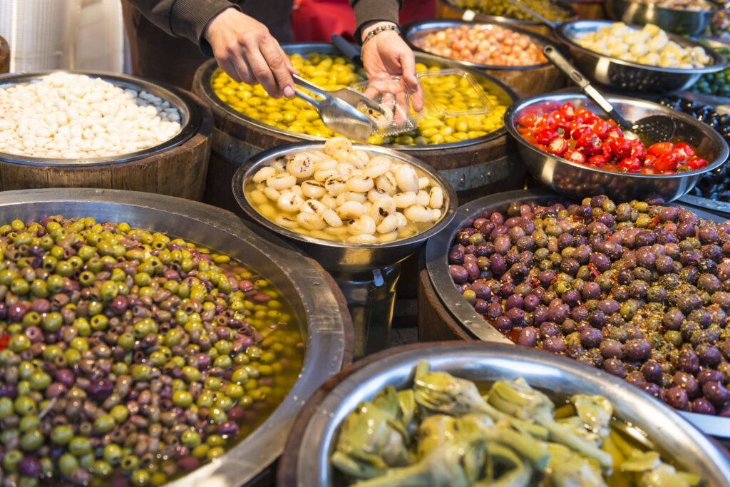 A market stall in Sicily displays a variety of olives, onions, tomatoes, and other pickled vegetables in large bowls. A person's hand is scooping pickled onions using a utensil. The colorful display features green, black, and purple olives in the foreground, showcasing vibrant flavors.