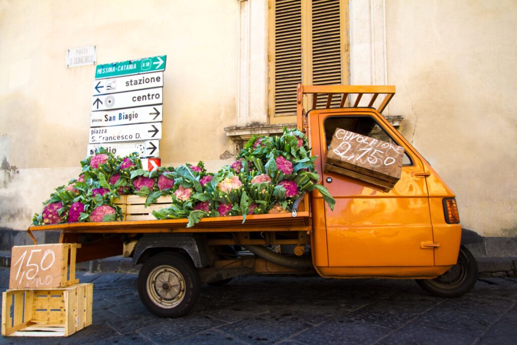 A small, orange, three-wheeled truck is parked on a street, loaded with crates of leafy green and purple vegetables. A handwritten sign on the truck reads "Formato 2,50" and there's another sign showing a price of "1,50". Directional street signs are visible in the background.