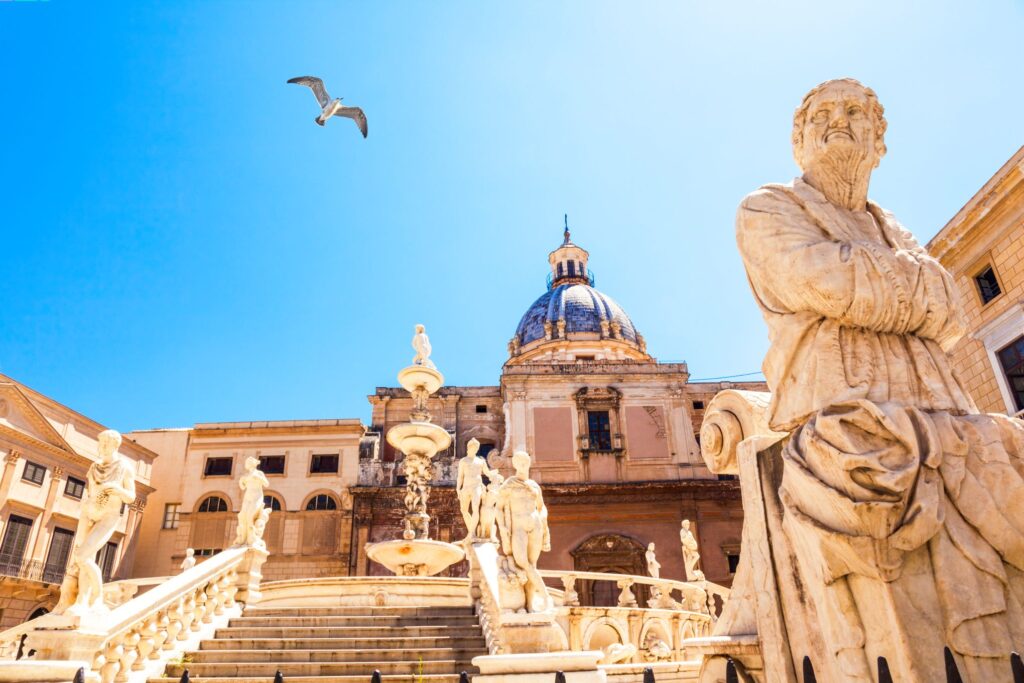 A historic plaza featuring ornate statues and a grand staircase leads to a building with a domed roof under a clear blue sky. A seagull is in flight above the scene, enhancing the serene atmosphere as it captures the timeless beauty that evokes flavors of Sicily.