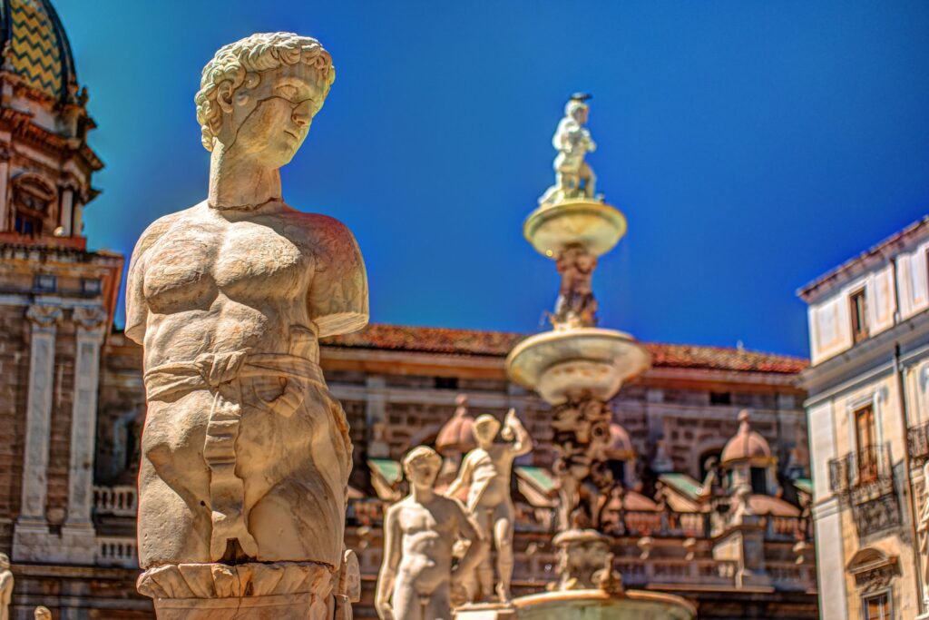 A close-up of a marble statue in a historical plaza under a clear blue sky. The statue depicts an ancient figure with detailed features. In the background, other statues and architectural elements of a grand building and a fountain evoke the rich cultural flavors of Sicily.