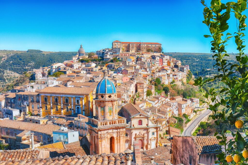 Panoramic view of a hillside town with colorful buildings and a prominent blue-domed church in the foreground. The town, reminiscent of Sicily's charm, is surrounded by lush green hills under a clear blue sky. Lemon branches frame the right side, hinting at the region's culinary flavors.