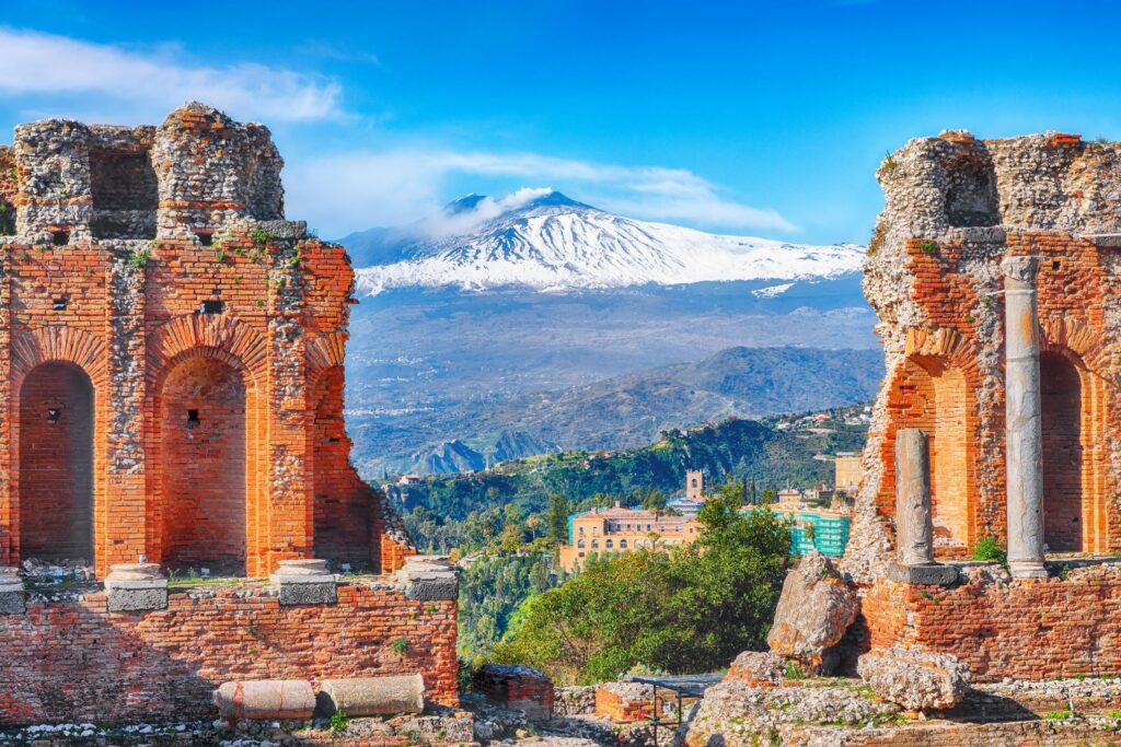 Ancient brick ruins frame a scenic view of a snow-capped mountain, surrounded by a lush green landscape under a clear blue sky, reminiscent of the rich flavors of Sicily.