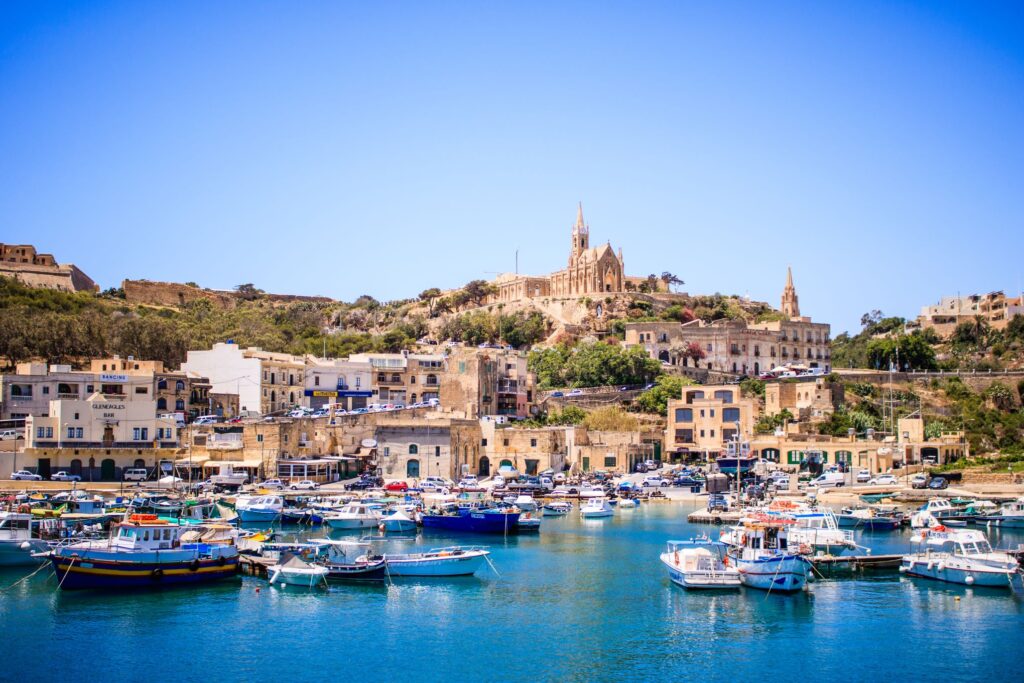 A picturesque coastal town featuring a serene harbor filled with various boats docked on crystal clear blue water. The town, reminiscent of the flavors of Sicily, is built on a hillside with historic stone buildings, and a prominent church with a tall spire stands at the top under a clear blue sky.