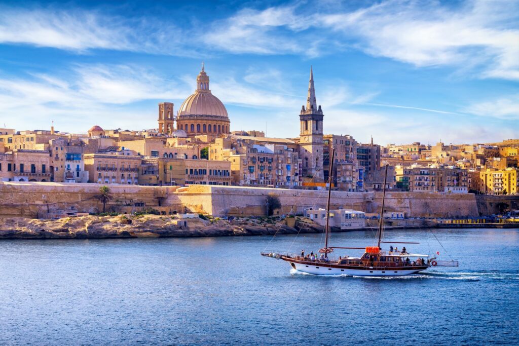 Historic cityscape of Valletta, Malta, featuring St. Paul's Cathedral and a fortified skyline against a blue sky with scattered clouds. A wooden sailing boat cruises the calm blue waters of the harbor in the foreground, evocative of flavors of Sicily blending into its rich maritime heritage.