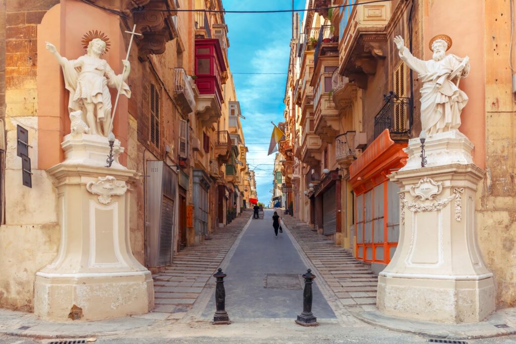 A narrow, picturesque street in an old European town reminiscent of Malta, featuring tall, ornate statues on either side of the entrance. The buildings are historic with balconies and worn facades. A few people are walking up the slightly inclined street under a clear, blue sky.