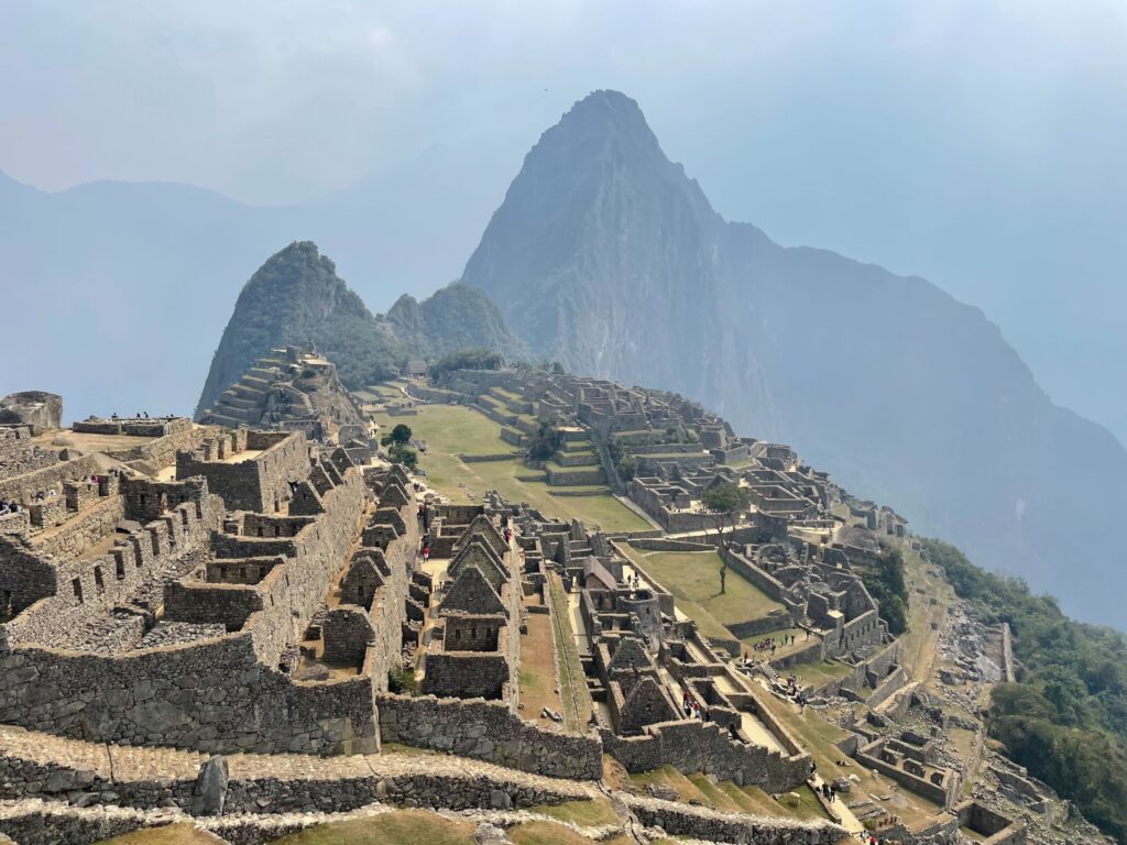 A breathtaking view of Machu Picchu in Peru, an ancient Incan city set high in the Andes Mountains. The image shows the ruins of stone buildings and terraces on a steep hillside, with the iconic Huayna Picchu peak rising dramatically in the background under a hazy sky.