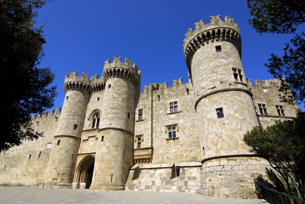 A historic stone castle with multiple cylindrical towers topped with battlements, set against a clear blue sky. The entrance features a large arched doorway, and the walls are adorned with small windows. Trees frame the scene on both sides, capturing the essence of a spectacular Greece adventure.