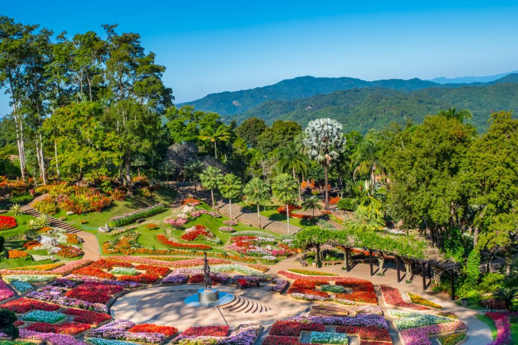 A vibrant botanical garden with diverse flower beds arranged in intricate patterns, numerous trees, a circular water feature at the center, and a hilly, forested landscape in the background under a clear blue sky – perfect for those exploring Thailand tour packages.