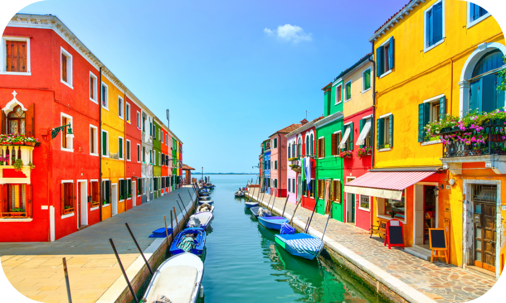 Colorful canal street in Burano, Italy, with vibrant red, yellow, and green buildings offering visual deals along a narrow waterway. Small boats are moored along the banks under a clear blue sky.