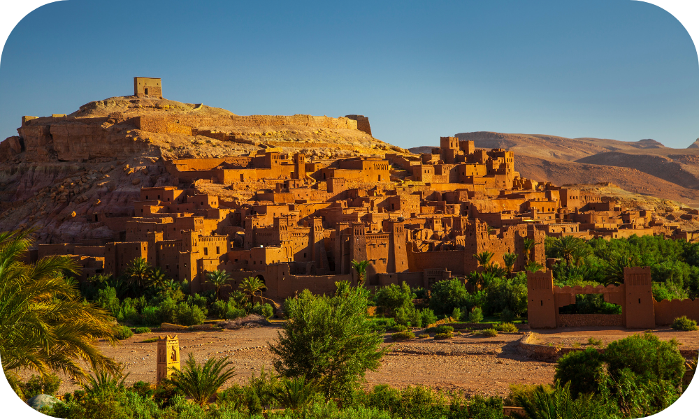 A majestic view of the ancient earthen buildings of Ait Benhaddou under a clear blue sky. The fortified village, often featured in travel deals, is surrounded by lush greenery and desert hills, showcasing its iconic reddish-brown architecture against the natural landscape.