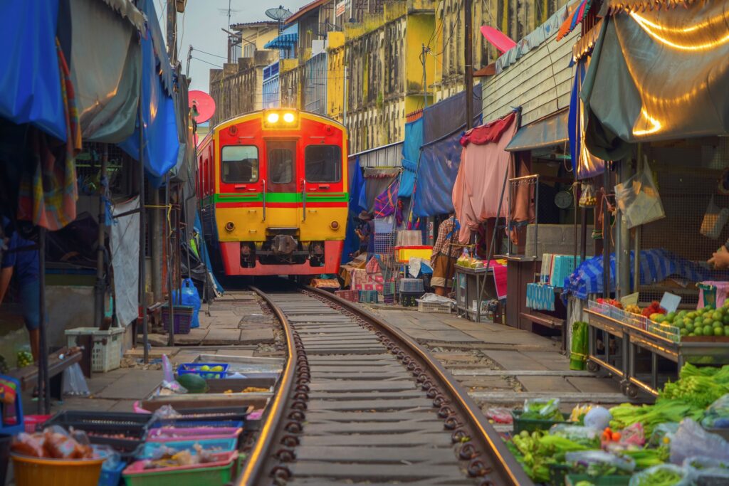 A vibrant train travels through a narrow market in Thailand, with stalls and colorful umbrellas tightly packed on either side of the railway tracks. Fresh produce, goods, and incredible deals are displayed on the ground and tables, making use of every inch of space around the tracks.