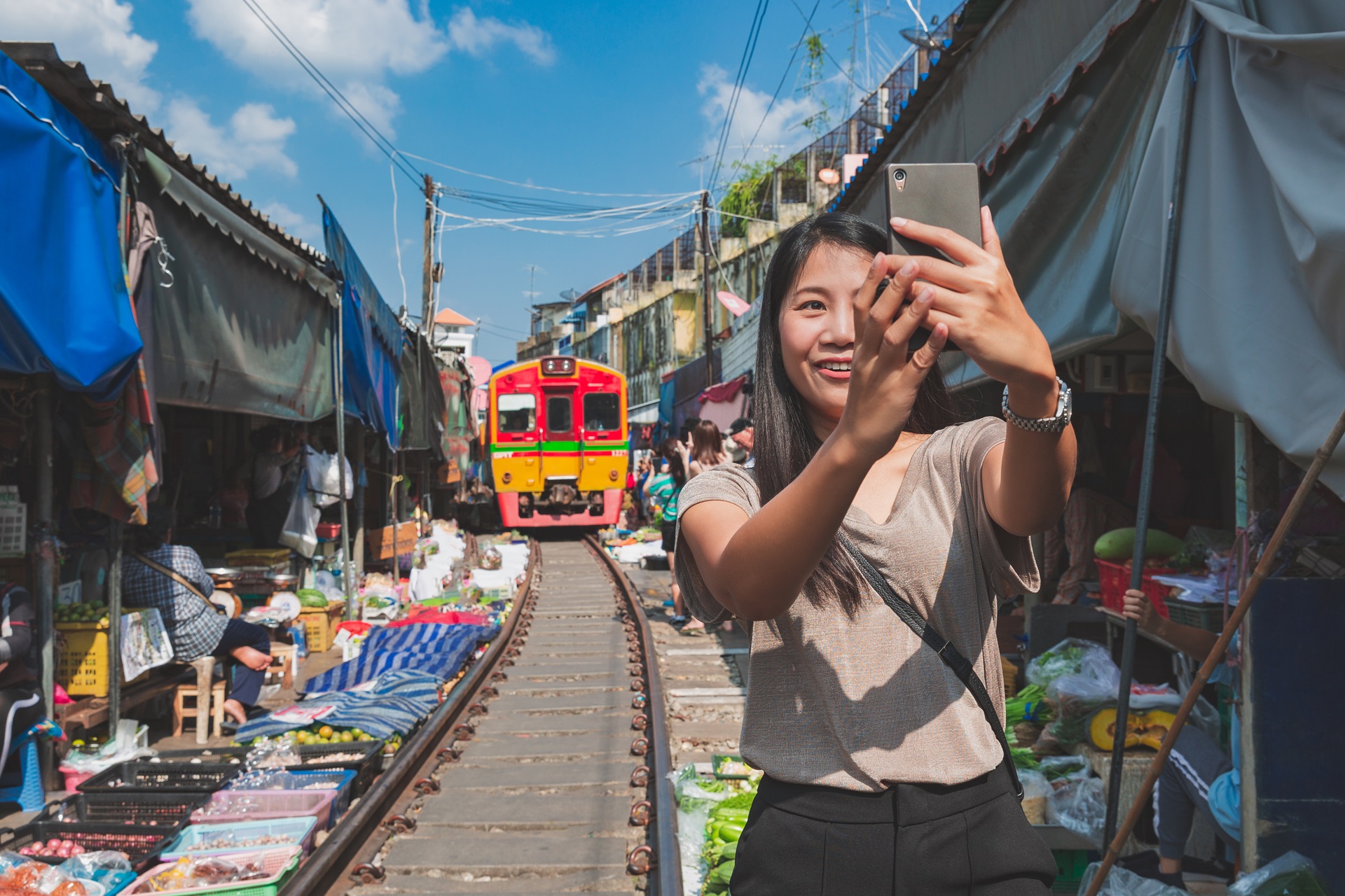 A woman takes a selfie while standing on railway tracks in a bustling market, famous for its amazing deals. Stalls with colorful goods and produce line both sides of the tracks. A red and yellow train approaches in the background under a clear blue sky.