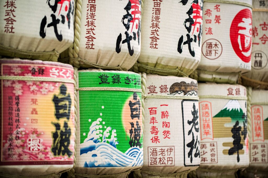 A close-up of rows of stacked traditional Japanese sake barrels, decorated with colorful and intricate kanji calligraphy, symbols, and nature scenes. Discover Japan through the barrels' unique designs, each showcasing different imagery and text in a vibrant display.