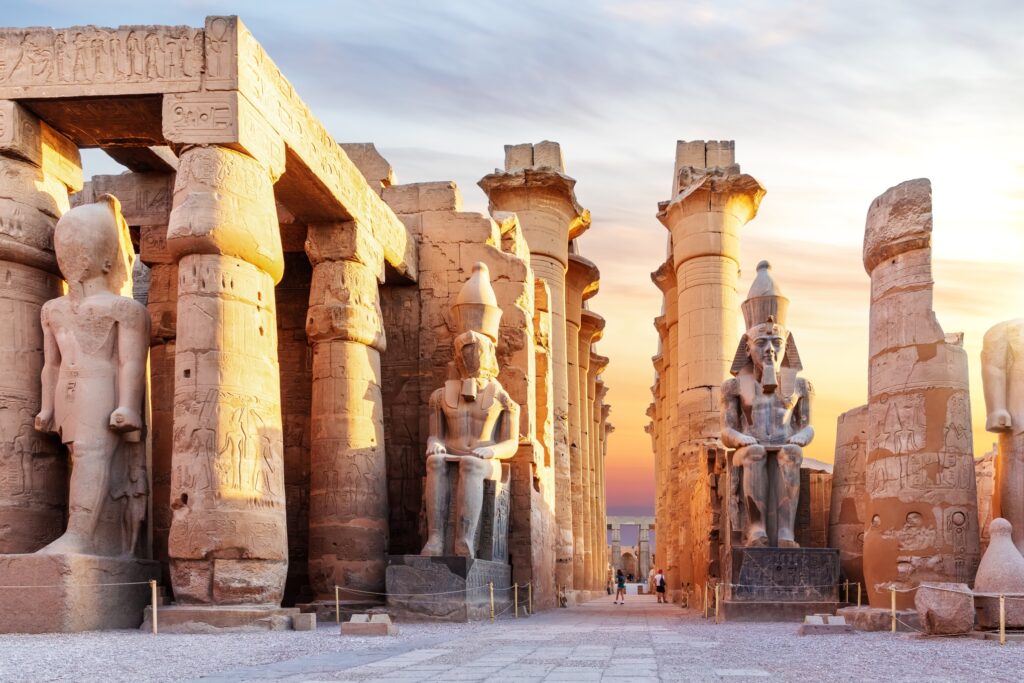 Ancient Egyptian temple ruins at sunset, featuring colossal statues and towering columns adorned with hieroglyphs. The path between the structures leads into the distance under a beautiful sky transitioning from orange to purple hues, inviting visitors to explore through curated Egypt tour packages.