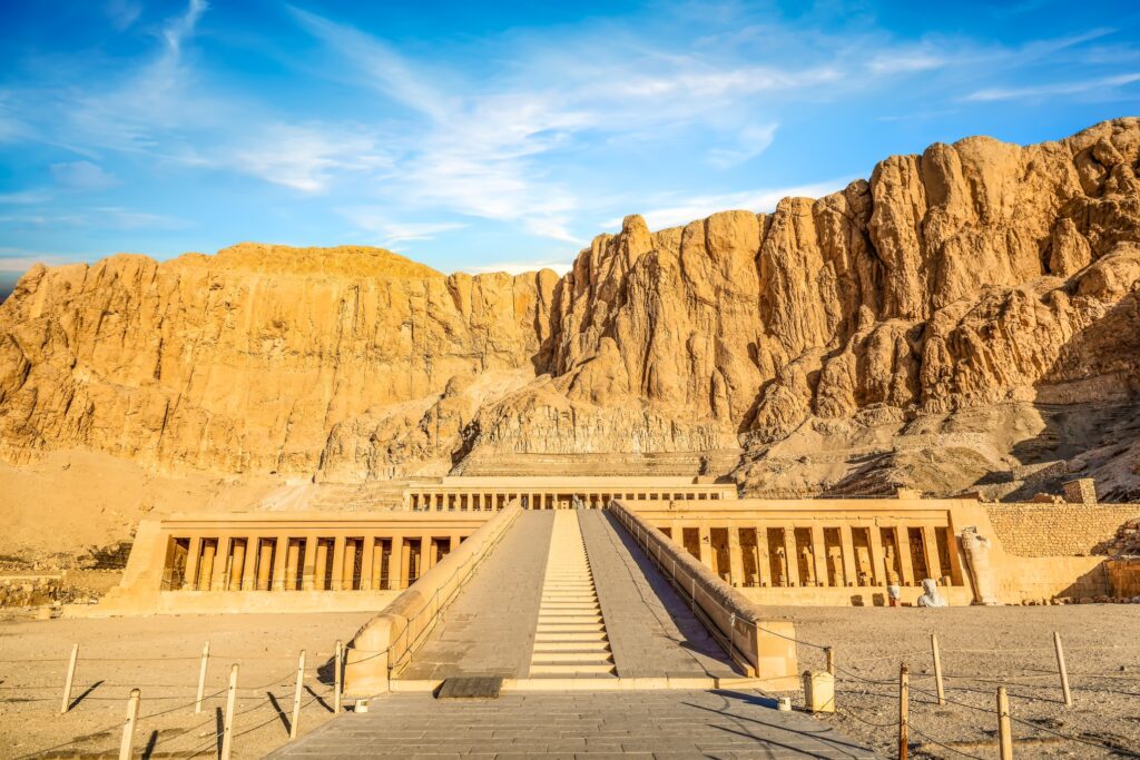 A wide-angle view of the Mortuary Temple of Hatshepsut in Deir el-Bahari, Egypt, showcases its grand, symmetrical structure with rows of columns, steps leading up to the temple, and rugged cliffs in the background under a clear blue sky—an essential highlight in egypt tour packages.