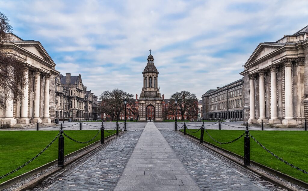 A wide walkway lined with cobblestone and black chains leads to a central bell tower with classical architecture. The path, flanked by green lawns and grand white-columned buildings under a partly cloudy sky, deals an air of scholarly elegance. The symmetrical scene is likely a university campus.