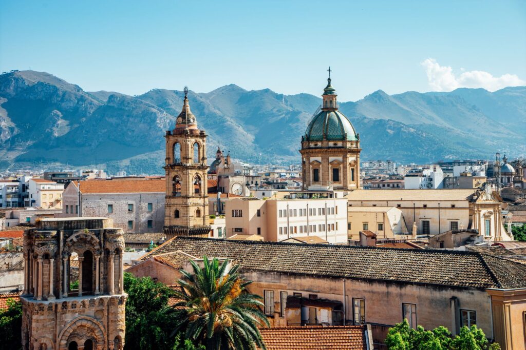 A vibrant cityscape view of Sicily featuring historic architecture with domed and bell towers set against a backdrop of rugged, green mountains on a clear day. Palm trees and rooftops populate the foreground, highlighting a blend of urban and natural beauty with the flavors of Mediterranean charm.