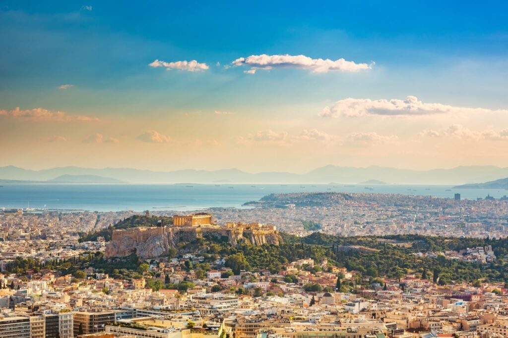 Aerial view of the Acropolis in Athens, Greece, bathed in sunlight with the ancient Parthenon visible on the rocky hill. The sprawling cityscape leads out to the sparkling blue sea and distant mountains under a partly cloudy sky. Spectacular Greece awaits you, perfect for an unforgettable Island Cruise.