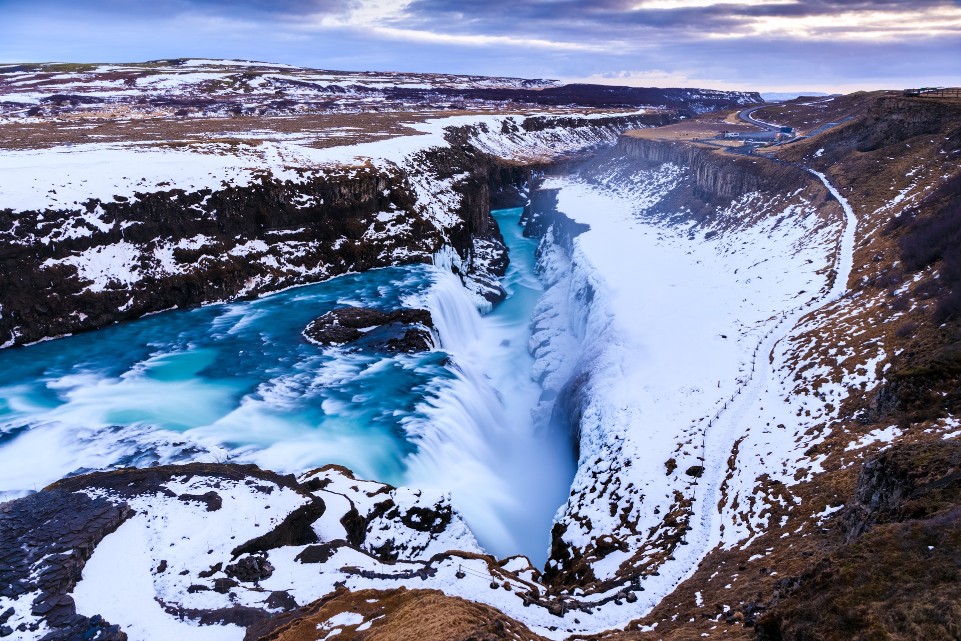 A breathtaking view of a powerful waterfall cascading into a deep canyon surrounded by snow-covered cliffs. The river flows through a rugged landscape with patches of snow and brown earth. The sky above is partly cloudy, adding a dramatic touch to the scene that nature's beauty offers without any deals required.