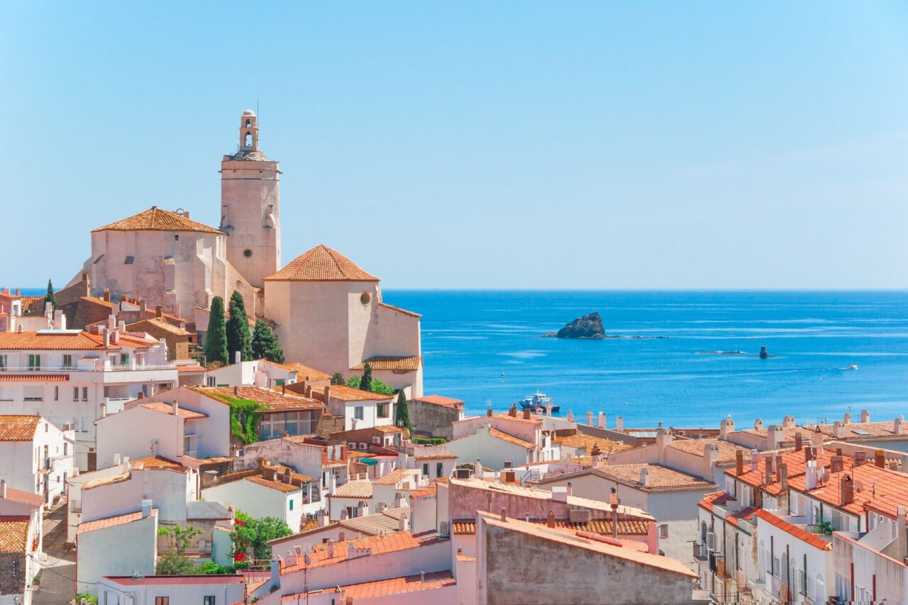 A picturesque coastal village with white houses and terracotta rooftops. A prominent church with a bell tower stands on a hill overlooking the vibrant blue sea. There's a small boat near the rocky outcrops in the water under a clear, bright sky—an ideal spot for the Best of Spain tour.