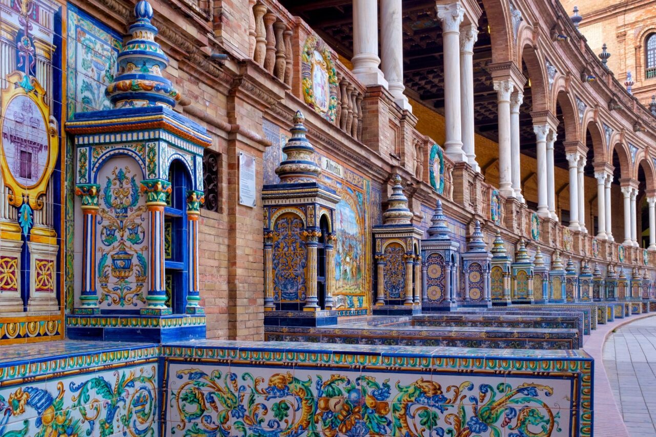 Intricate, colorful ceramic tiles and elaborate columns line the curved wall of Plaza de España in Seville, Spain. Each tile section depicts unique, detailed artwork, showcasing rich patterns and designs beneath classic archways.