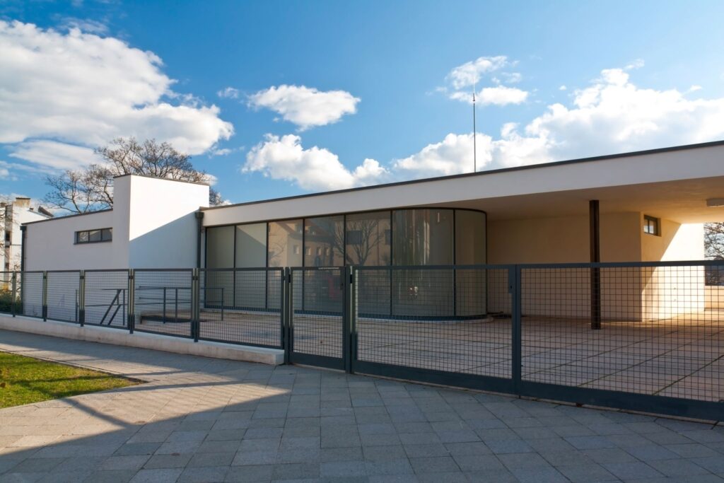 Modern, single-story building with large glass windows and a curved wall section, surrounded by a metal fence. The exterior features a minimalist design with a flat roof, reminiscent of the sleek architecture seen during a Prague Vienna Budapest tour. The sky above is blue with scattered clouds. A paved walkway leads to the entrance.