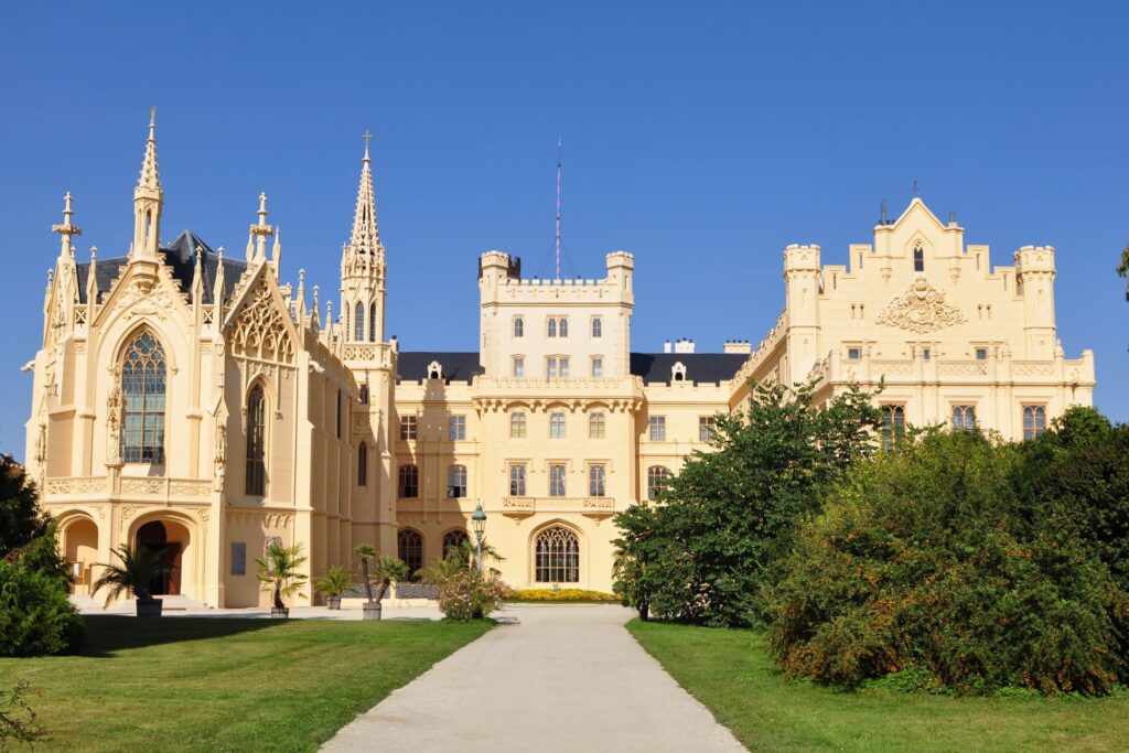 A large historic castle featuring Gothic and Renaissance architectural styles, with tall spires, arched windows, and an intricate facade. The building is set against a clear blue sky, surrounded by lush green trees and a manicured lawn with a gravel pathway—perfect for any Prague or Vienna tour.