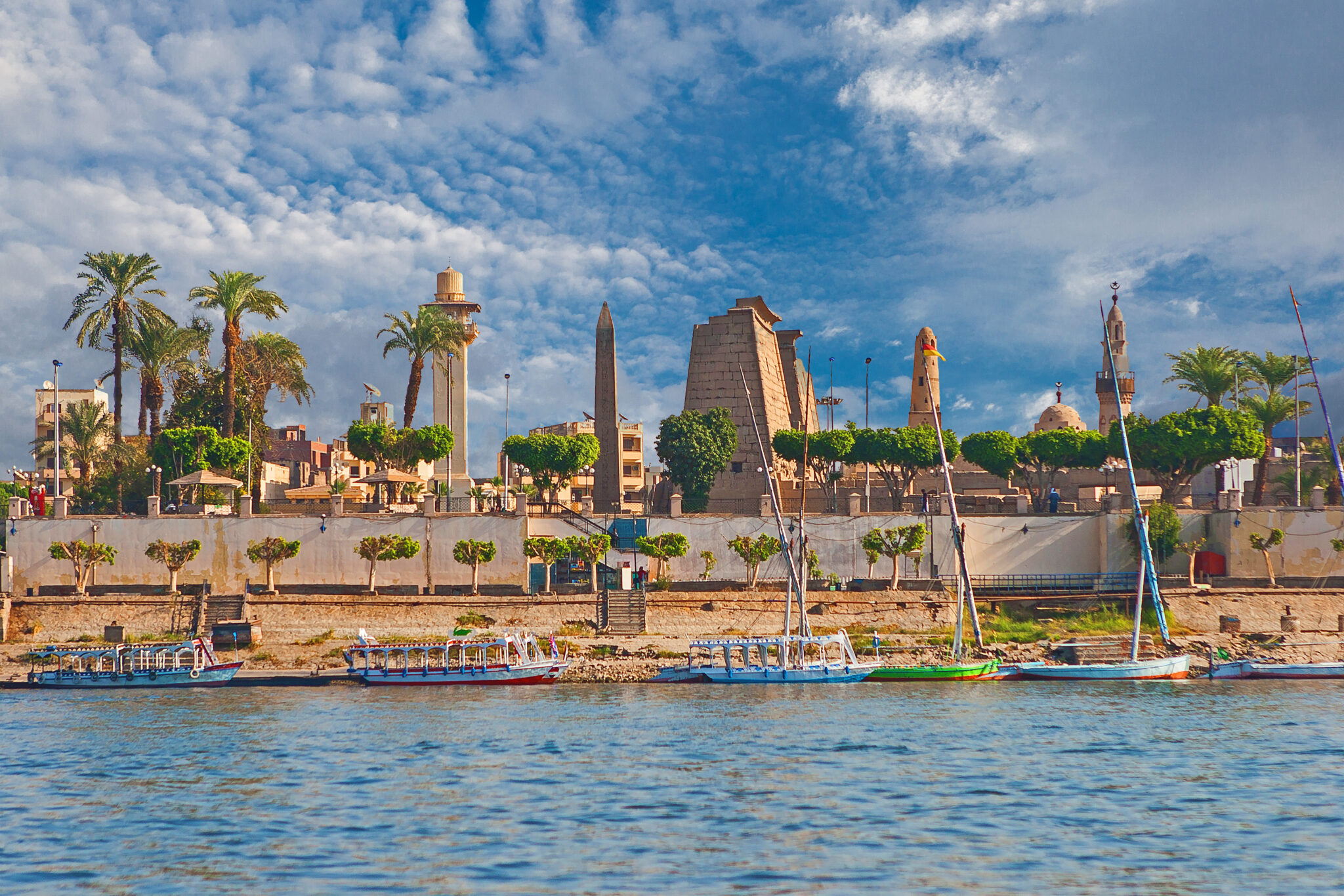 A view of Luxor Temple in Egypt from across the Nile River. The temple, palm trees, and various ancient structures are visible against a backdrop of a partly cloudy sky. Several traditional boats are moored along the river's edge in the foreground—perfect for bucket-list experiences with friends.