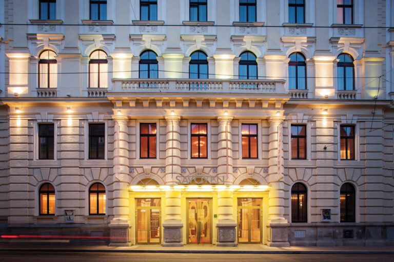 The image shows a grand, multi-story historical building with intricate architectural details, reminiscent of the charming scenes you’d find on a Prague or Vienna tour. The facade features columns, arched windows, and soft lighting that highlights its elegant design. The entrance has glass doors illuminated by warm lights, creating a welcoming glow.