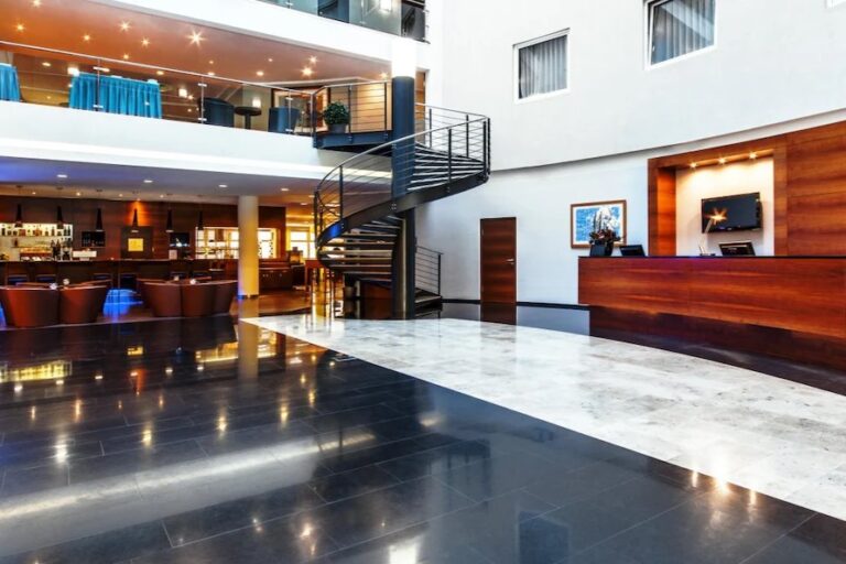 A modern hotel lobby with a shiny black and white marble floor, a spiral staircase leading to an upper level with seating areas, a reception desk on the right with a flat-screen TV, and a bar in the background. Warm lighting adds an inviting atmosphere perfect for guests on their Prague-Vienna-Budapest tour.