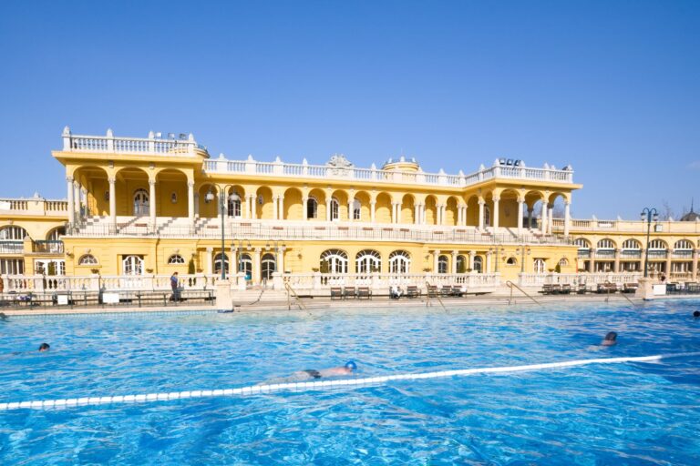 A vibrant yellow historic building with arched windows and balconies overlooks a large outdoor swimming pool. A few swimmers are enjoying the pool under a clear blue sky, highlighting a sunny day. The opulent, classical architecture offers a taste of iconic cities like Prague or Vienna.