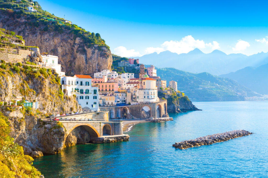 A picturesque coastal town with colorful buildings perched on steep cliffs overlooking a calm blue sea offers the perfect setting for bucket-list experiences with friends. The town features arched bridges and terraces, while lush green mountains rise under a clear blue sky. A rock jetty extends into the sea.