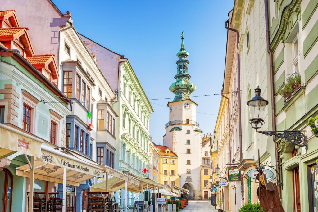 A picturesque European street with pastel-colored buildings, outdoor café seating, and a prominent historic tower with a green spire in the background under a clear blue sky. Reminiscent of Prague or Vienna, the street is lined with charming shops and architectural details perfect for any tour.