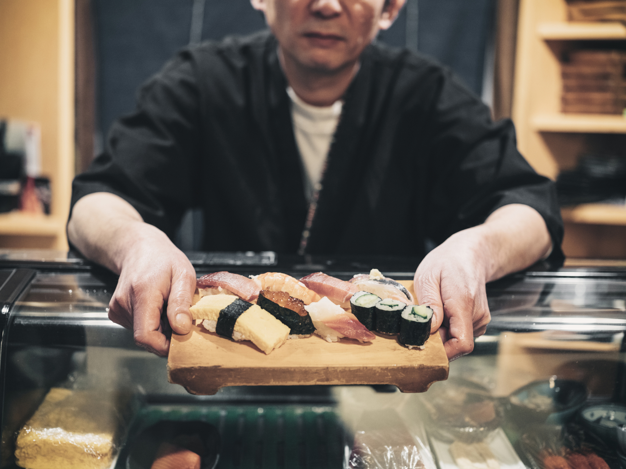 A chef in a black uniform holds a wooden tray with an assortment of sushi over a glass counter. The tray features various types of sushi, including nigiri, maki, and sashimi. In the background—a blurred interior of what appears to be a sushi restaurant—awaits your next bucket-list experience with friends.