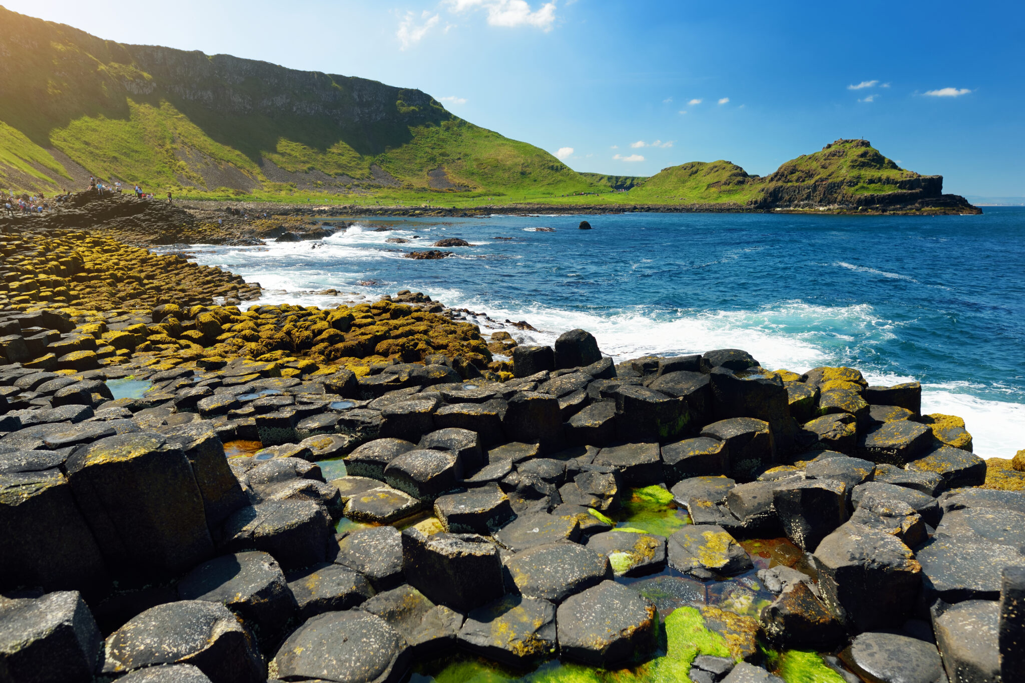 A view of the Giant's Causeway in Northern Ireland, featuring interlocking basalt columns formed from ancient volcanic activity. The columns line the shore next to a blue sea with waves and a green, hilly landscape under a bright, sunny sky—perfect for bucket-list experiences with friends.