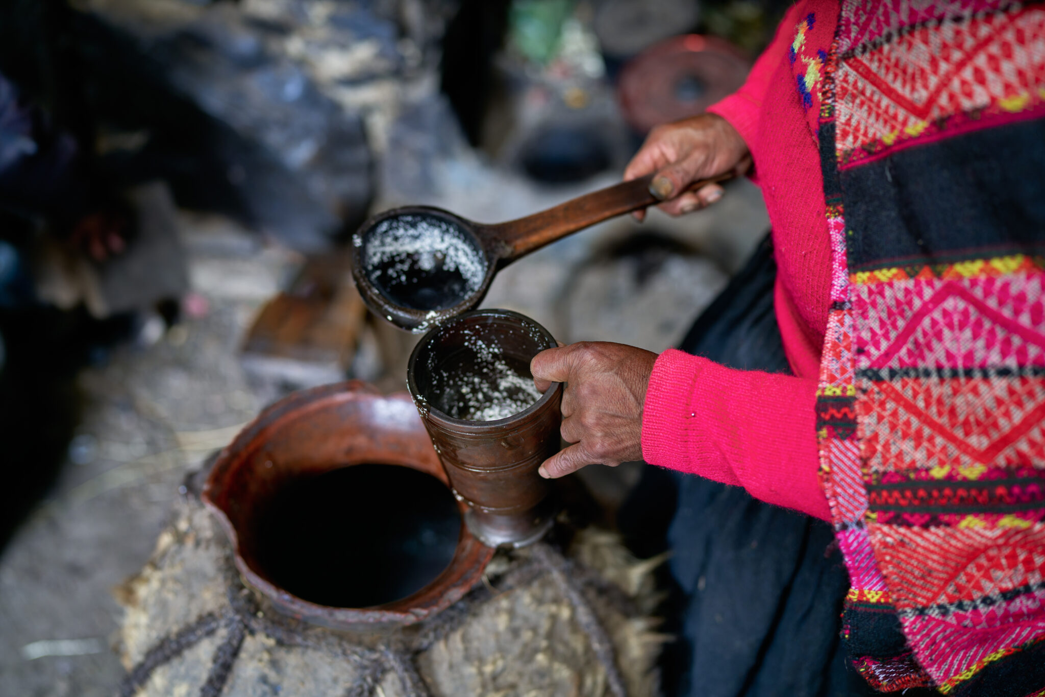 A person wearing a red garment with colorful patterns is pouring a drink into a wooden cup from a gourd-shaped container using a ladle. The scene appears to be indoors, highlighting hands and the drink being poured—a perfect snapshot of bucket-list experiences with friends.