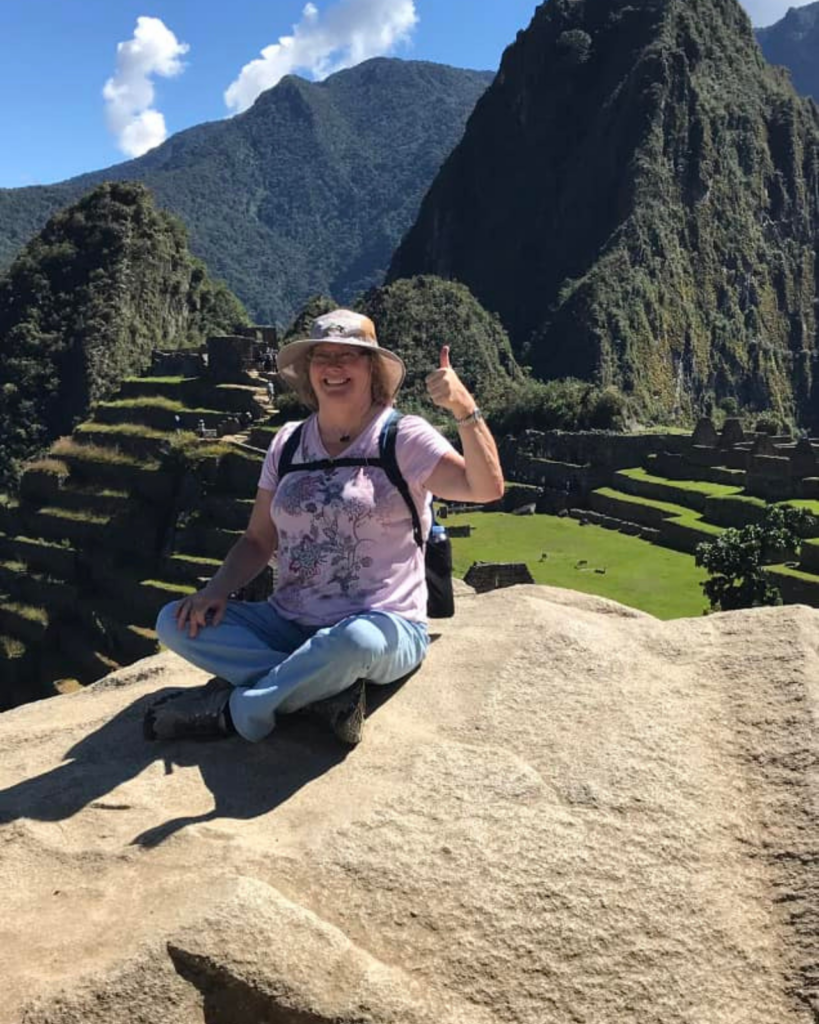 A solo person wearing a sunhat, glasses, a light pink T-shirt, and light blue pants is sitting on a rock with a large mountain and ancient ruins in the background. They are smiling and giving a thumbs-up gesture with their right hand, clearly thrilled about finding great travel deals. The sky is clear and sunny.