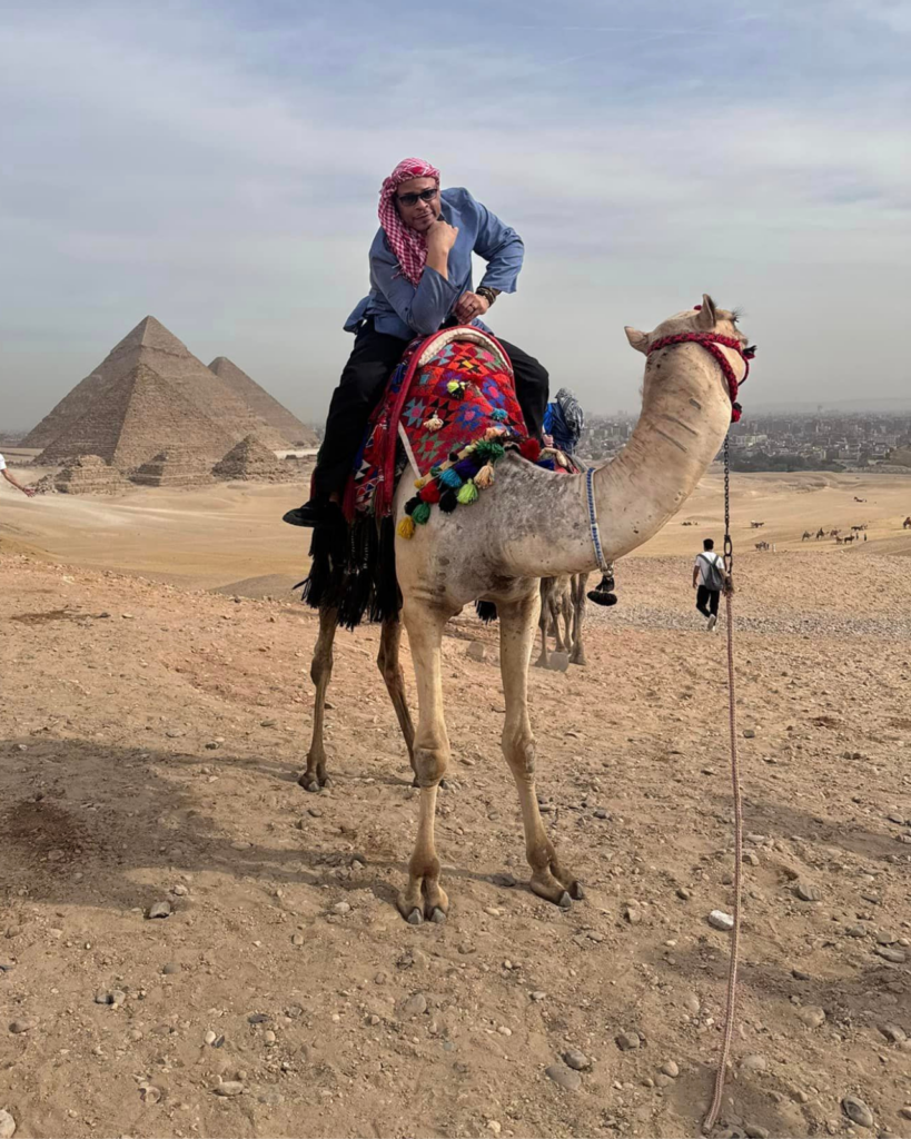 A solo person wearing a blue jacket and a red and white headscarf poses thoughtfully while sitting on a decorated camel. In the background, the Pyramids of Giza stand under a clear sky, reminding travelers of timeless adventures and ancient deals carved in history.