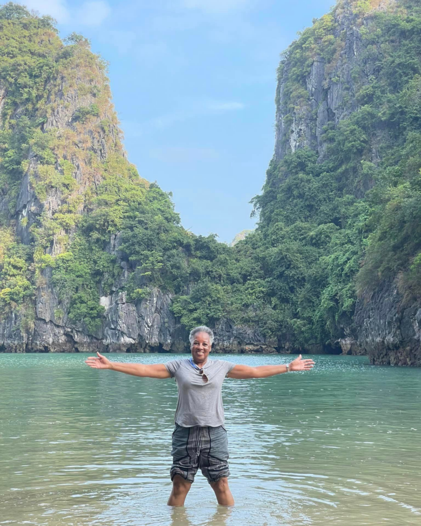 A solo person standing knee-deep in clear, turquoise water, arms outstretched, in a picturesque cove with steep, lush green cliffs on either side. The sky is clear blue, and the serene scene is as inviting as the best travel deals. The person is smiling and wearing a light grey t-shirt and shorts.