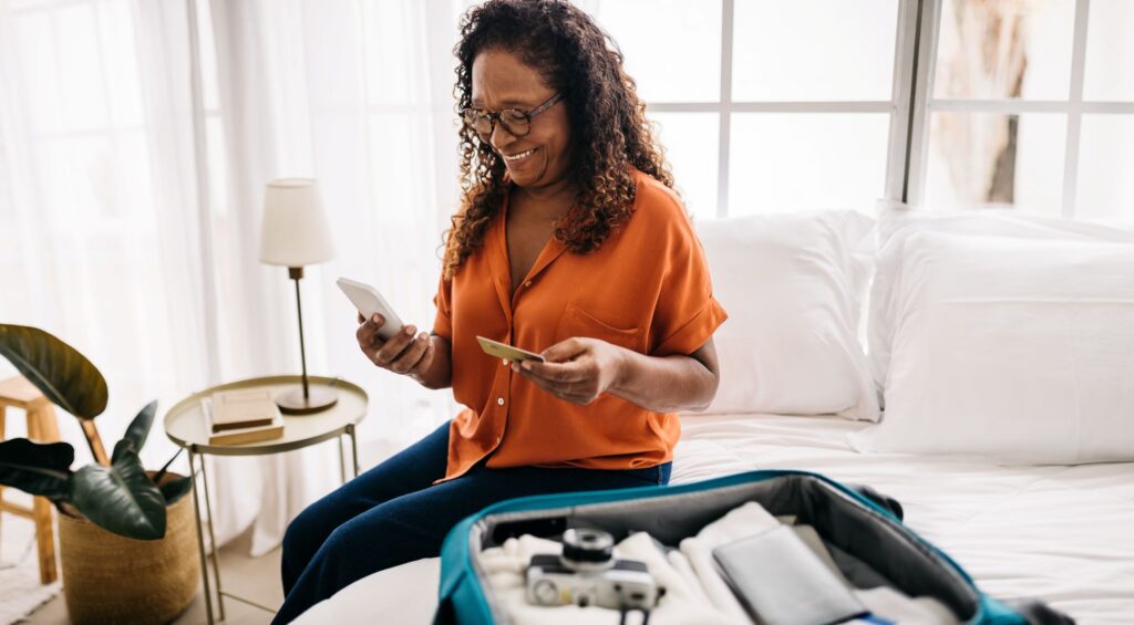 A person with curly hair and glasses sits on a bed holding a smartphone and a credit card, smiling. An open suitcase with clothes and a camera can be seen on the bed beside them. The room has a soft, natural light with a plant and a lamp in the background.